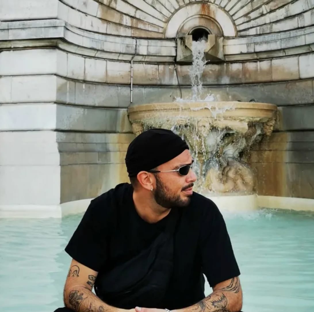 Zutzut sitting by a stone fountain, wearing sunglasses and a black head wrap